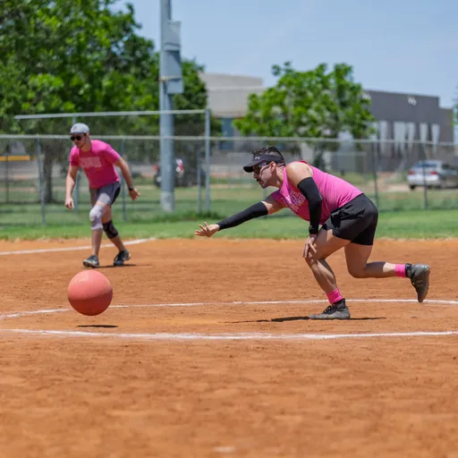 A player in a pink shirt and arm sleeves is rolling a kickball on a dirt field, while another player in similar attire stands ready behind them; the setting is an outdoor kickball field with fencing, green trees, and buildings in the background.