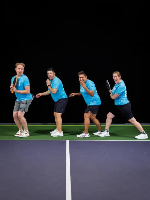 Four pickleball players in matching blue shirts pose playfully in a line on an indoor court, each holding a pickleball paddle and appearing to be mid-action or mid-laugh. The court lines and green playing surface are visible, and the background is dark, highlighting the players' energetic expressions and team spirit.
