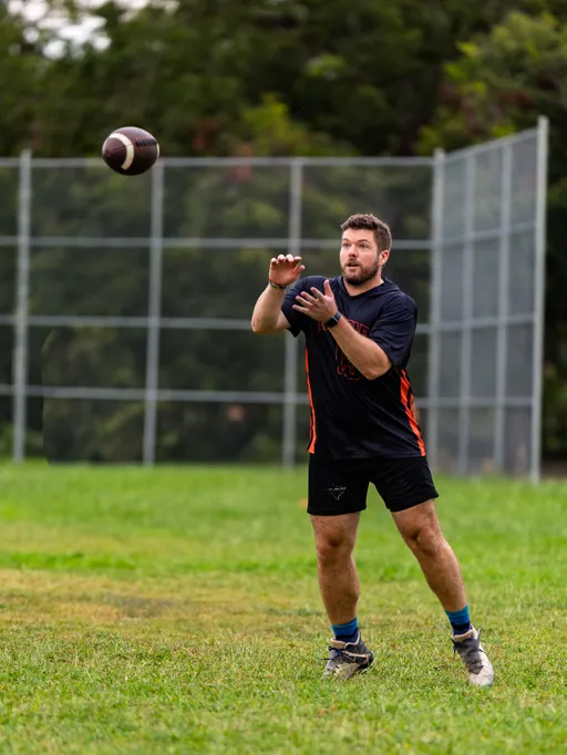 An ATX Heat (Orange) player raises their hands to catch a football during Gay Bowl 2025 practice on a grassy park field, wearing a black jersey with orange side panels, black shorts, blue socks, and cleats, with a chain-link fence and trees in the background.