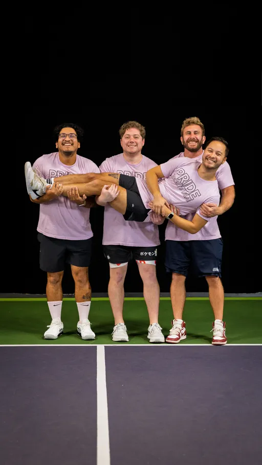 Four pickleball players stand on an indoor court, wearing matching "Pride" t-shirts, with three players holding a fourth teammate horizontally while everyone smiles and poses for the photo.