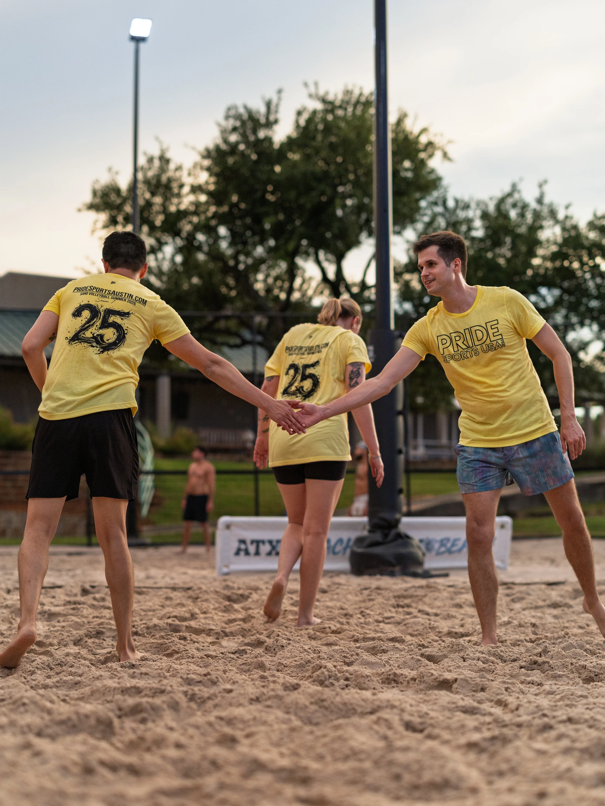 Three volleyball players wearing matching yellow "Pride Sand Volleyball" shirts are on a sand court, with two of them giving each other a high five and another walking nearby; the scene takes place outdoors with sports equipment and a fence visible in the background.