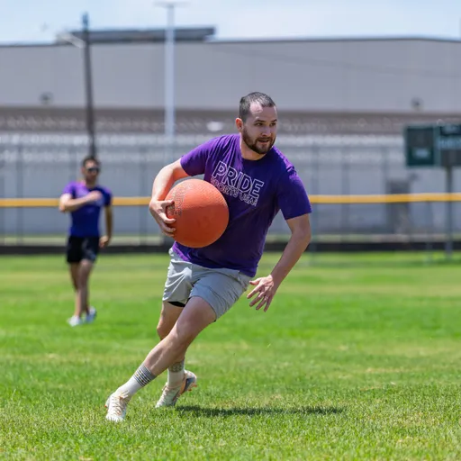 A player wearing a purple "Pride Kickball" shirt and athletic shorts is running across a grassy kickball field while holding a kickball, with another player visible in the background.