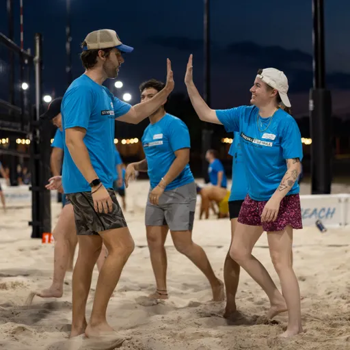 A group of Stonewall sand volleyball players in matching blue shirts gathers by the net at dusk, exchanging high-fives after a point. The lit sand court shows boundary lines and net posts; players wear shorts, some with caps, patterned shorts, and name tags.