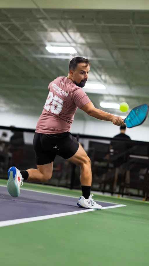 A pickleball player in a maroon jersey with the number 25 and black shorts lunges to reach a ball with a blue paddle on an indoor court. The setting features a green and blue playing surface with visible court boundaries and other players or spectators in the background.