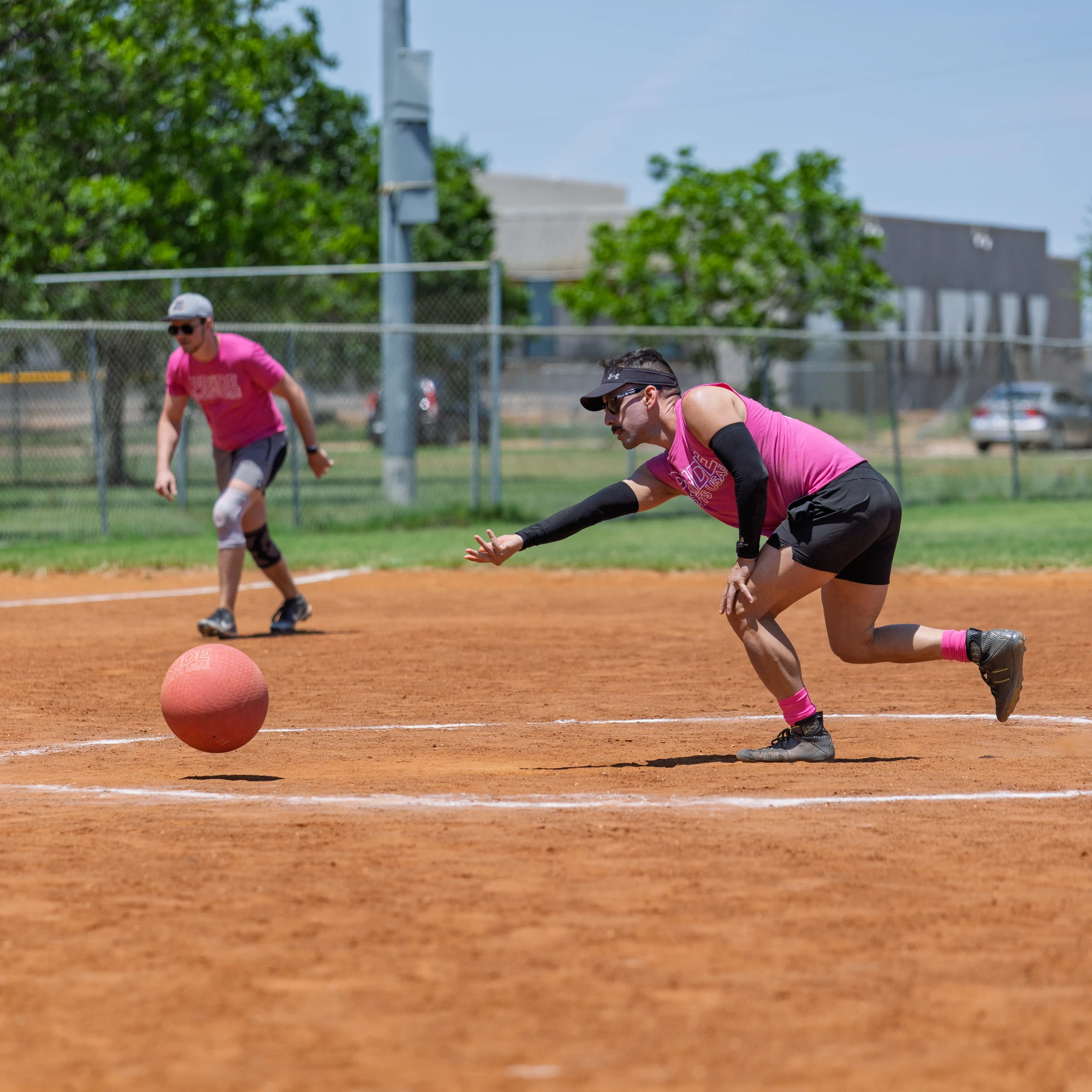 A player in a pink shirt and arm sleeves is rolling a kickball on a dirt field, while another player in similar attire stands ready behind them; the setting is an outdoor kickball field with fencing, green trees, and buildings in the background.