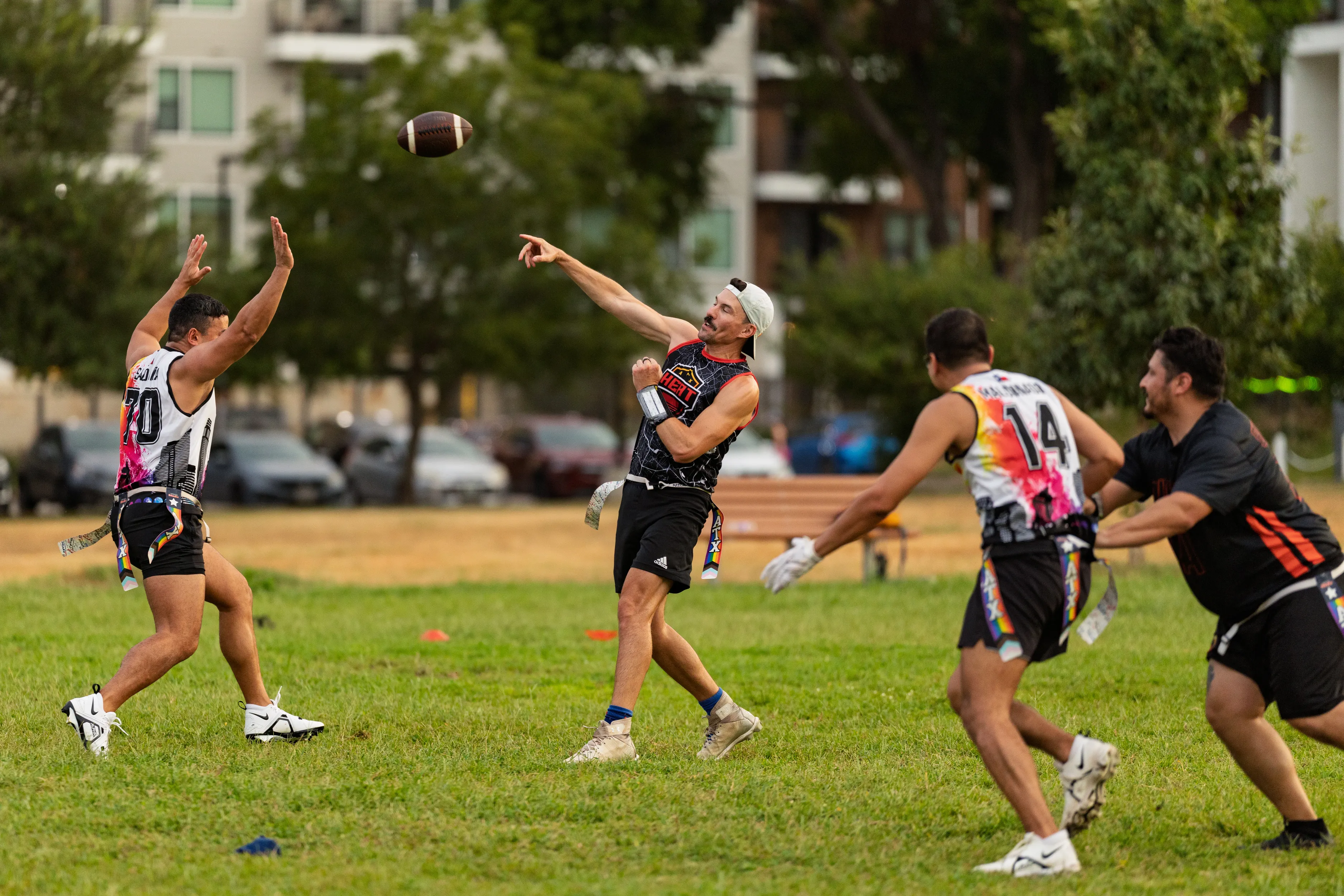 ATX Heat (Orange) players scrimmage in a city park: a quarterback in a black jersey with multicolor accents and a backward cap throws a pass as a receiver reaches up and two defenders close in; all wear numbered jerseys, flag belts, and cleats, with cones marking the field.