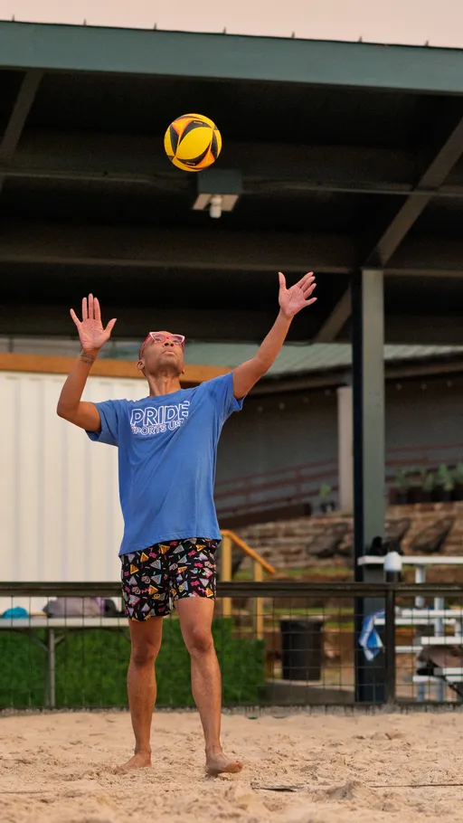 A volleyball player wearing a blue "PRIDE SPORTS USA" shirt, colorful shorts, and sunglasses is about to serve the ball on a sand volleyball court. The environment includes a covered area and outdoor seating in the background.