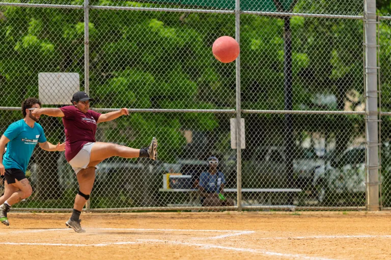 A player in a maroon shirt and black cap is kicking a red kickball on a dirt field, while a competitor in a teal shirt runs behind them; a chain-link fence and green trees are visible in the background.
