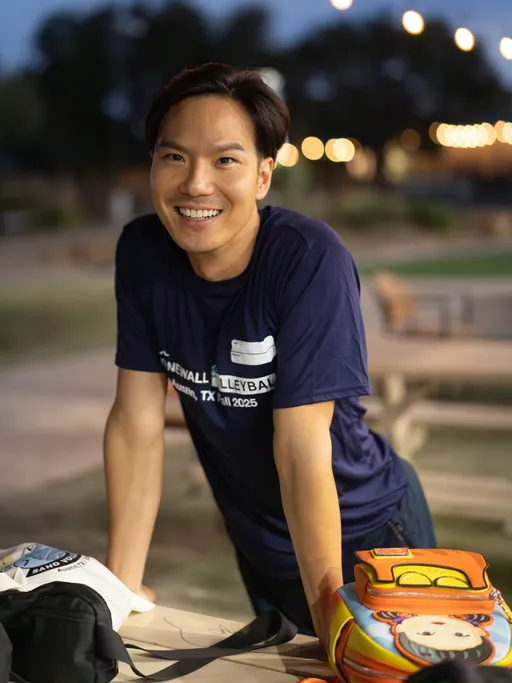 A player in a navy Stonewall Volleyball Austin, TX Fall 2025 shirt leans on a picnic table, smiling during a break at dusk near the sand courts. A tote and a colorful backpack with a cartoon face rest on the table, with blurred park lights, benches, and the outdoor volleyball area in the background.