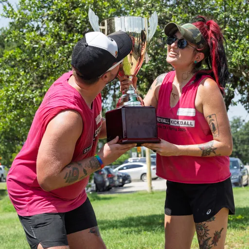 Two players wearing matching pink "Stonewall Kickball" jerseys and athletic gear are holding up a large trophy outdoors, smiling and celebrating on a sunny day with trees and parked cars in the background.