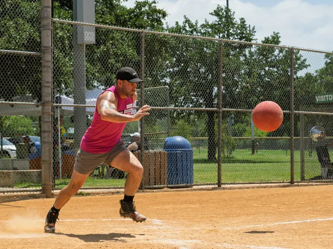 A kickball player wearing a pink sleeveless shirt, black cap, shorts, and athletic shoes is running on a dirt field, reaching forward with one arm toward a red kickball in midair near home plate. A chain link fence and green park area are visible in the background.