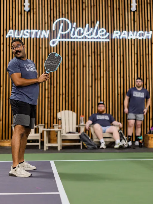 A player on a pickleball court prepares to swing a paddle, wearing athletic shorts, a "Pride Pickleball" t-shirt, and sneakers; in the background, two other participants in matching t-shirts observe from near wooden lounge chairs at the Austin Pickle Ranch indoor facility.