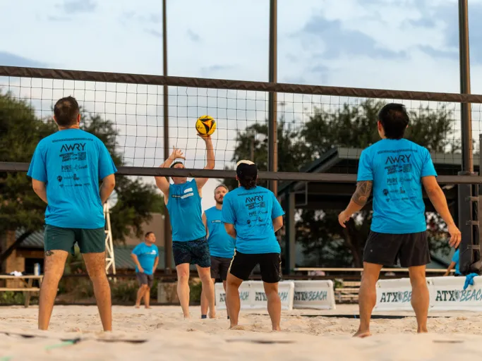 During a Stonewall Sand Volleyball match at an outdoor sand court, a player in a bright blue team shirt jumps at the net to set or block a yellow-and-black volleyball while teammates in matching shirts and dark shorts—one wearing a cap—wait in ready positions. Evening light, court banners, and a pavilion with trees frame the action.