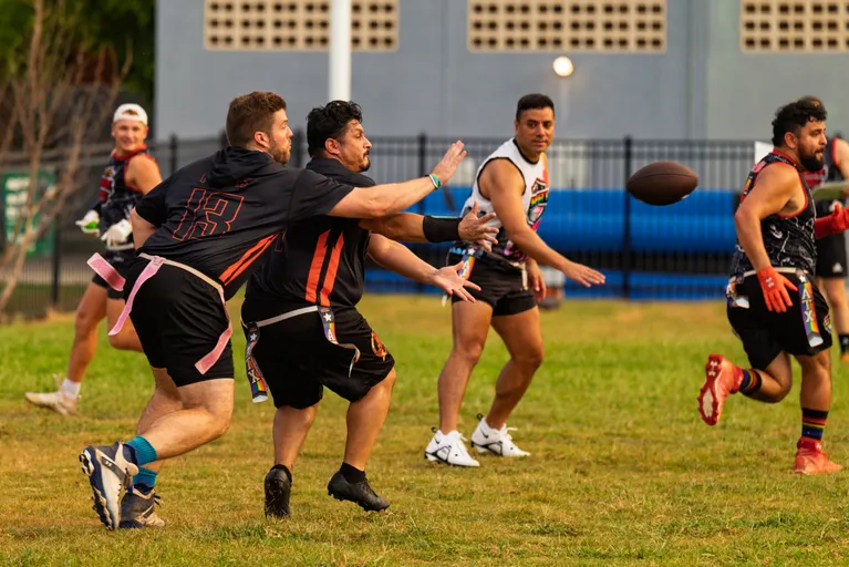 During a Gay Bowl 2025 practice, ATX Heat (Orange) athletes run a flag‑football drill on a grassy field: a short pass flies across the middle as a receiver reaches for it while a defender lunges to grab the flag belt. Players wear black-and-orange jerseys with pink flag belts, shorts, cleats, and some gloves, with a fence and building in the background.
