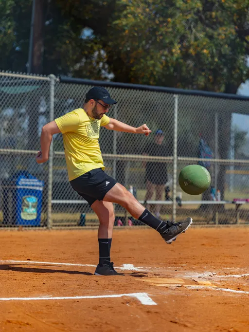 An athlete in a yellow team shirt kicks a green kickball off home plate, the ball rising midair from a dusty kickball field. The player wears a black cap, black shorts, knee-high socks, and cleats, with a chain-link backstop, benches, and trees behind.
