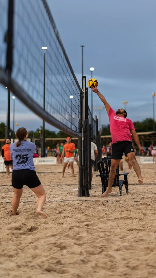 A sand volleyball player in a pink shirt leaps to block or spike the ball at the net, while a competitor in a gray shirt with the number 25 stands ready on the opposite side; the action takes place on an outdoor sand court surrounded by other players and illuminated lights.