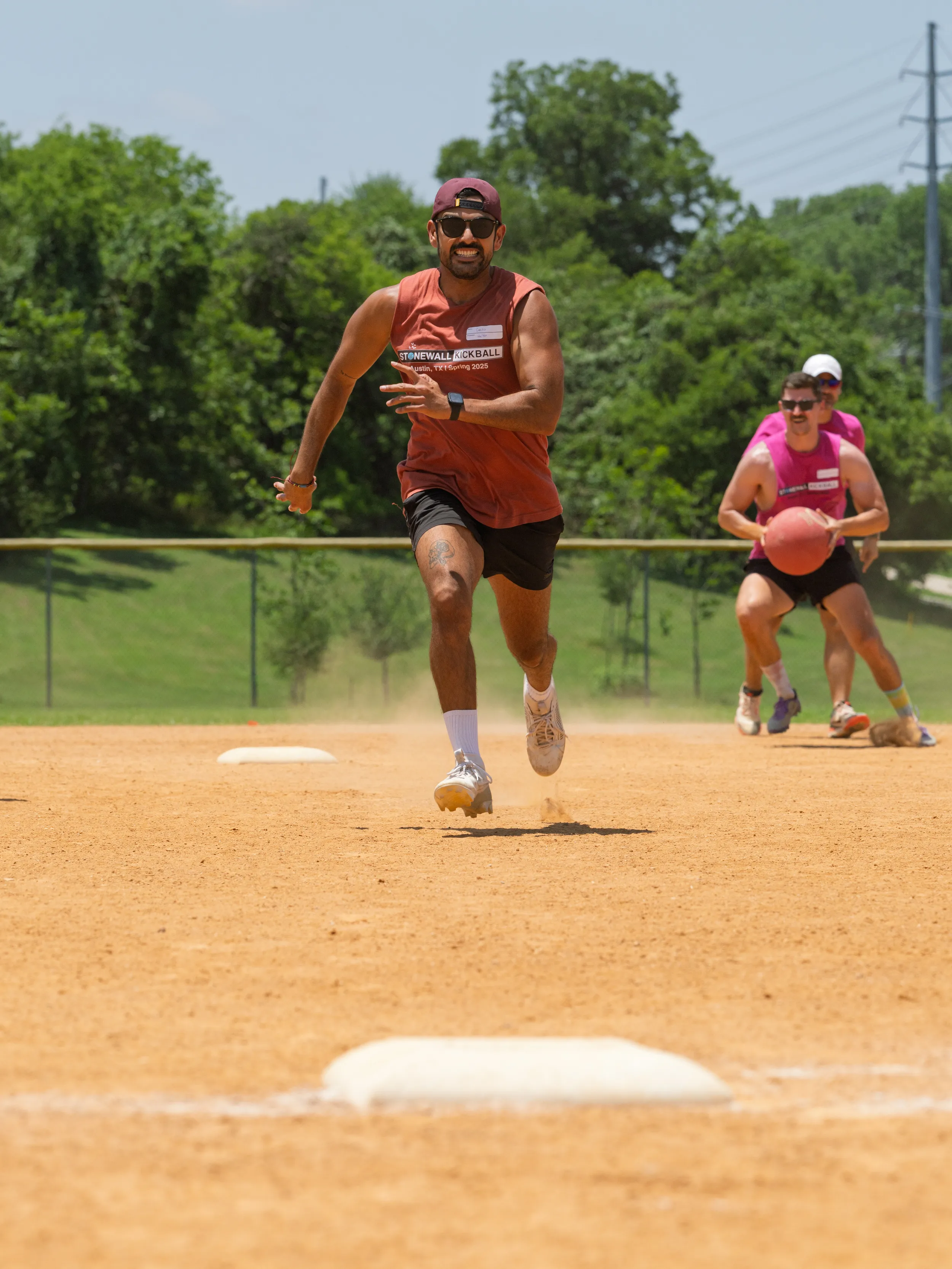 On a sunny kickball field, a runner in a maroon sleeveless jersey, black shorts, sunglasses, and a backward cap sprints toward a base while a defender in a pink jersey chases with the kickball in hand, kicking up dust across the infield with green trees beyond the fence.
