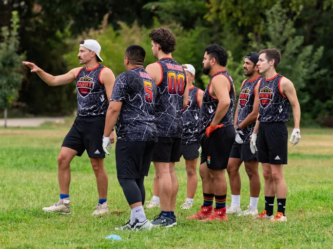 A group of ATX Heat (Orange) flag football players stands in a semicircle on a grassy field during Gay Bowl 2025 practice, with one player at left gesturing and giving instructions while the rest listen. They wear black, marble-pattern sleeveless jerseys with orange trim and “HEAT” crests and numbers, shorts, cleats, and receiver gloves; some have caps, rainbow socks, and armbands, with trees lining the background.