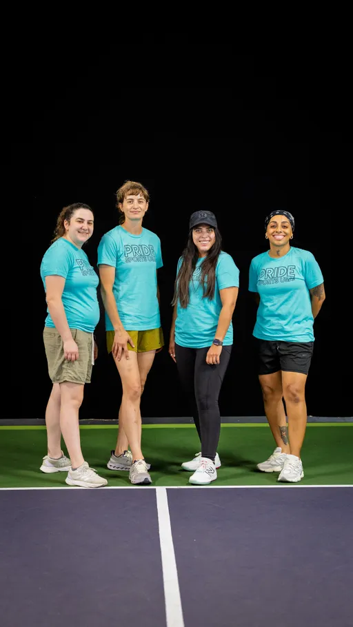 Four pickleball players standing together on an indoor pickleball court, all wearing matching blue "Pride Pickleball Summer 2025" shirts, athletic shorts or pants, and sneakers.