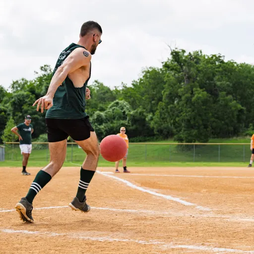 A player wearing a dark sleeveless shirt, shorts, striped socks, and athletic shoes is kicking a red kickball on a dirt field while teammates and other participants watch from the background. Trees and a fence line the edge of the kickball field.