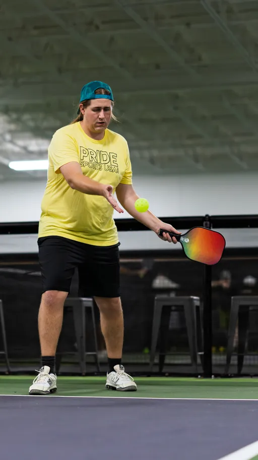 A player wearing a yellow "Pride Sports USA" shirt, black shorts, white athletic shoes, and a blue backwards cap is focused on serving a pickleball with a paddle on an indoor court.