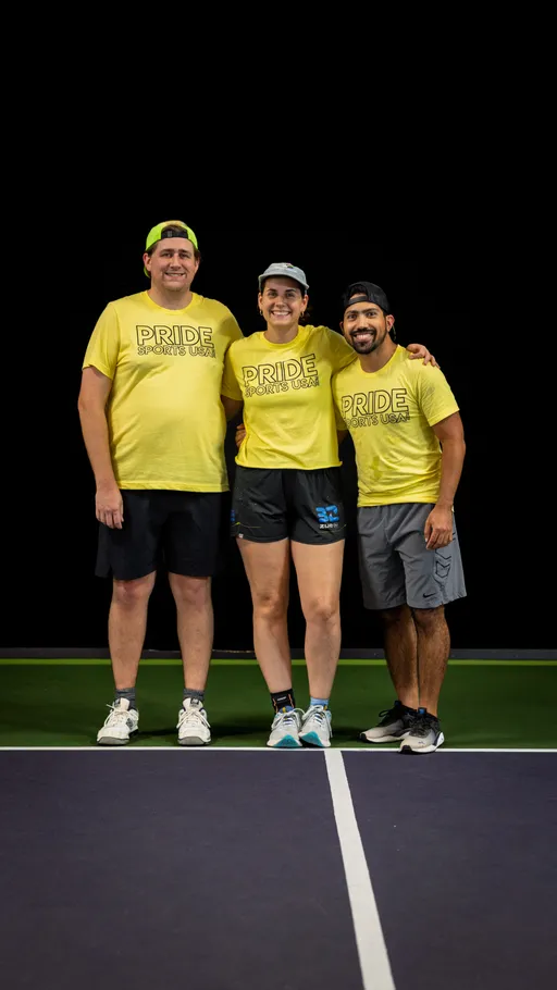 Three pickleball players stand side by side on an indoor court, each wearing matching yellow “Pride Sports USA” t-shirts, shorts, athletic shoes, and caps. The group is smiling and posing together in front of a dark background.