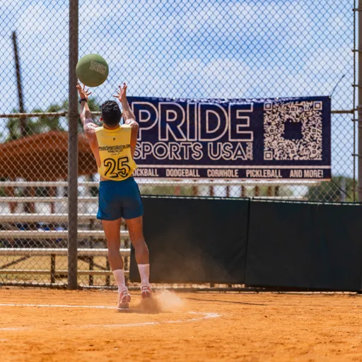 On a dusty kickball field, a Pitch Please player in a yellow jersey with number 25, blue shorts, and knee-high socks jumps at the backstop with arms raised to catch a green kickball, kicking up dust. A chain-link fence and a large PRIDE SPORTS USA banner fill the background in bright daylight.