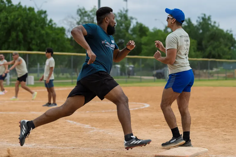 A kickball player in a dark shirt and shorts is sprinting toward a base while another player, wearing a light shirt, blue shorts, and a blue cap, stands ready at the base on a dirt kickball field with other athletes visible in the background.