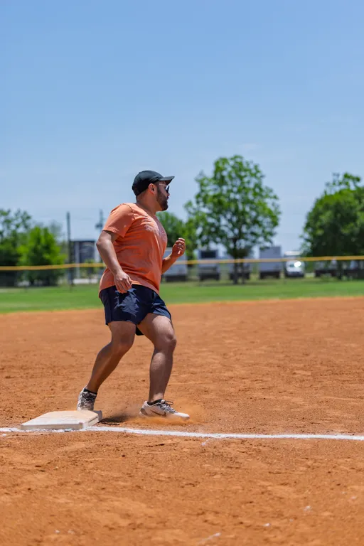A player wearing an orange shirt, dark shorts, sneakers, and a cap is running past a base on a kickball field under a clear sky, with grass and trees in the background.