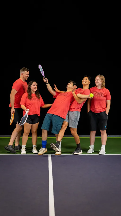 A group of five pickleball players wearing matching red "Pride" shirts stand together on an indoor court, with some holding paddles and one holding a pickleball, while laughing and posing playfully.