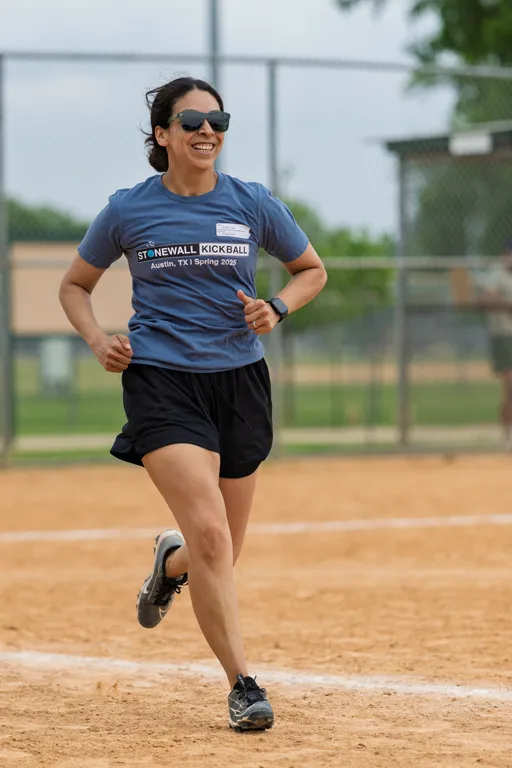 A player wearing sunglasses, a "Stonewall Kickball Austin, TX | Spring 2025" t-shirt, black shorts, and sports shoes is running on a kickball field, smiling during the game.