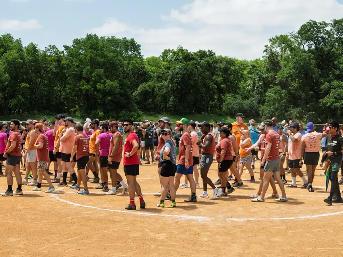A large group of kickball players stands and walks across a dirt kickball field, many wearing matching team shirts, athletic shorts, and various hats or sunglasses, with trees and a green landscape in the background.