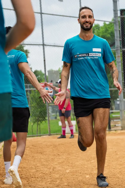 A kickball player in a blue Stonewall Kickball Spring 2025 shirt and black shorts runs on a dirt field, reaching out to high-five a teammate while others stand nearby behind a chain-link fence.