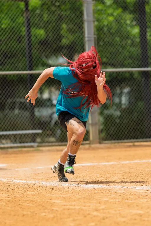 A player with long red hair is sprinting on a kickball field, wearing a teal team shirt, black shorts, and athletic shoes, with a tattoo visible on their left leg. The scene takes place on a dirt field with a chain-link fence and greenery in the background.