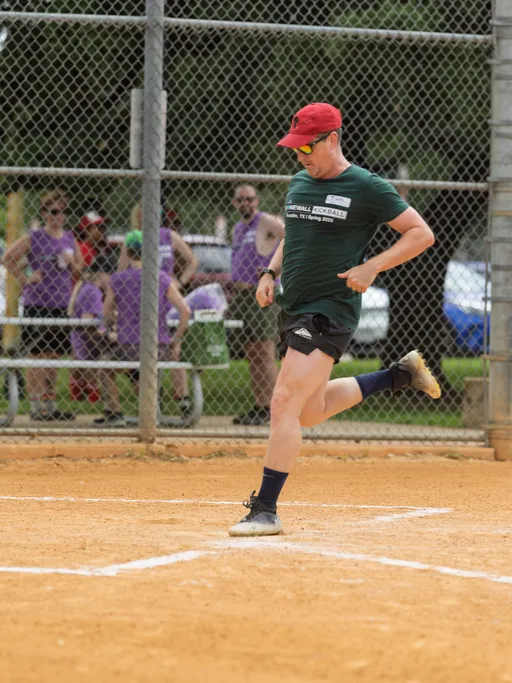 A player wearing a green Stonewall Kickball shirt, black shorts, a red cap, and athletic shoes is kicking a kickball on a sandy kickball field, with a chain-link fence and several people in purple shirts watching in the background.