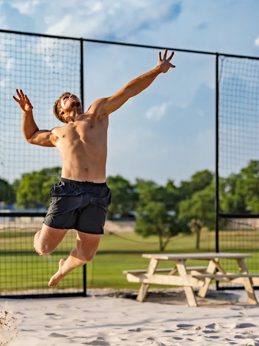 A volleyball player in black athletic shorts is leaping into the air on a sandy court, extending one arm overhead to make a play, with a net and outdoor park setting visible in the background.