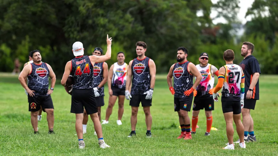 ATX Heat (Orange) players huddle on a grassy park field for Gay Bowl 2025 practice, listening as a teammate with an arm raised gives instructions. The team wears black jerseys with orange accents and the HEAT logo, shorts, cleats, and gloves; several have bright socks or rainbow sleeves, with cones marking the drill area and trees in the background.