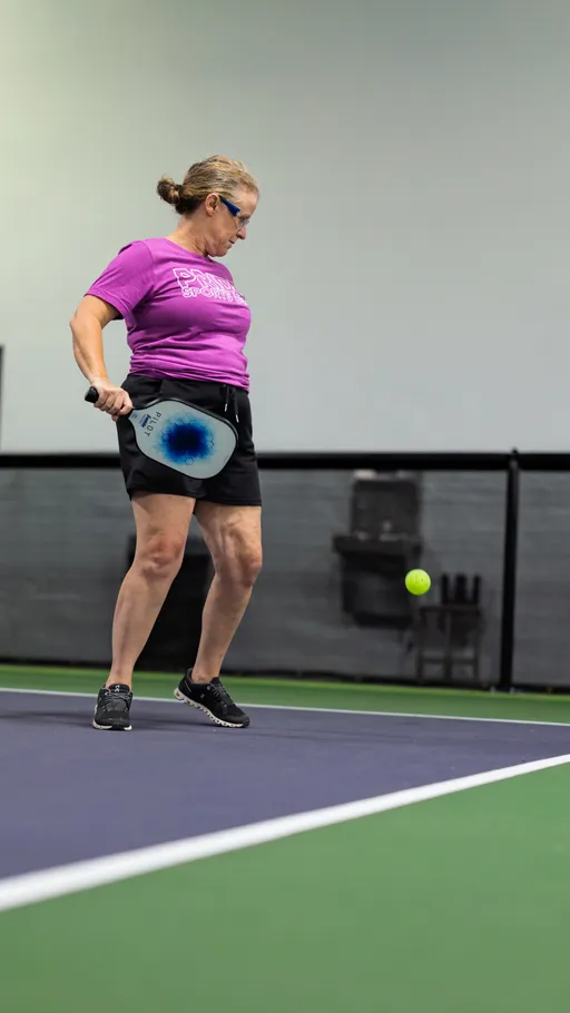 A pickleball player in a bright purple shirt and black shorts prepares to hit a ball with a paddle on an indoor court. The player is focused on the ball, which is in mid-air close to the ground.