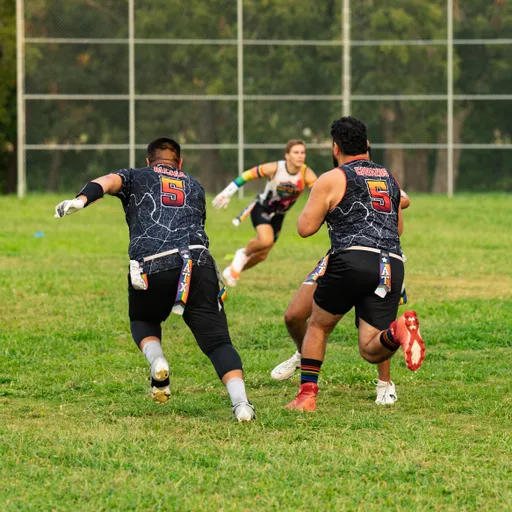On a grassy park field, ATX Heat (Orange) players run a Gay Bowl practice drill: two defenders in black patterned jerseys with red 5s, cleats, and multicolor flag belts close in as a ball carrier in similar gear sprints forward with the football; a chain-link fence and trees frame the background.