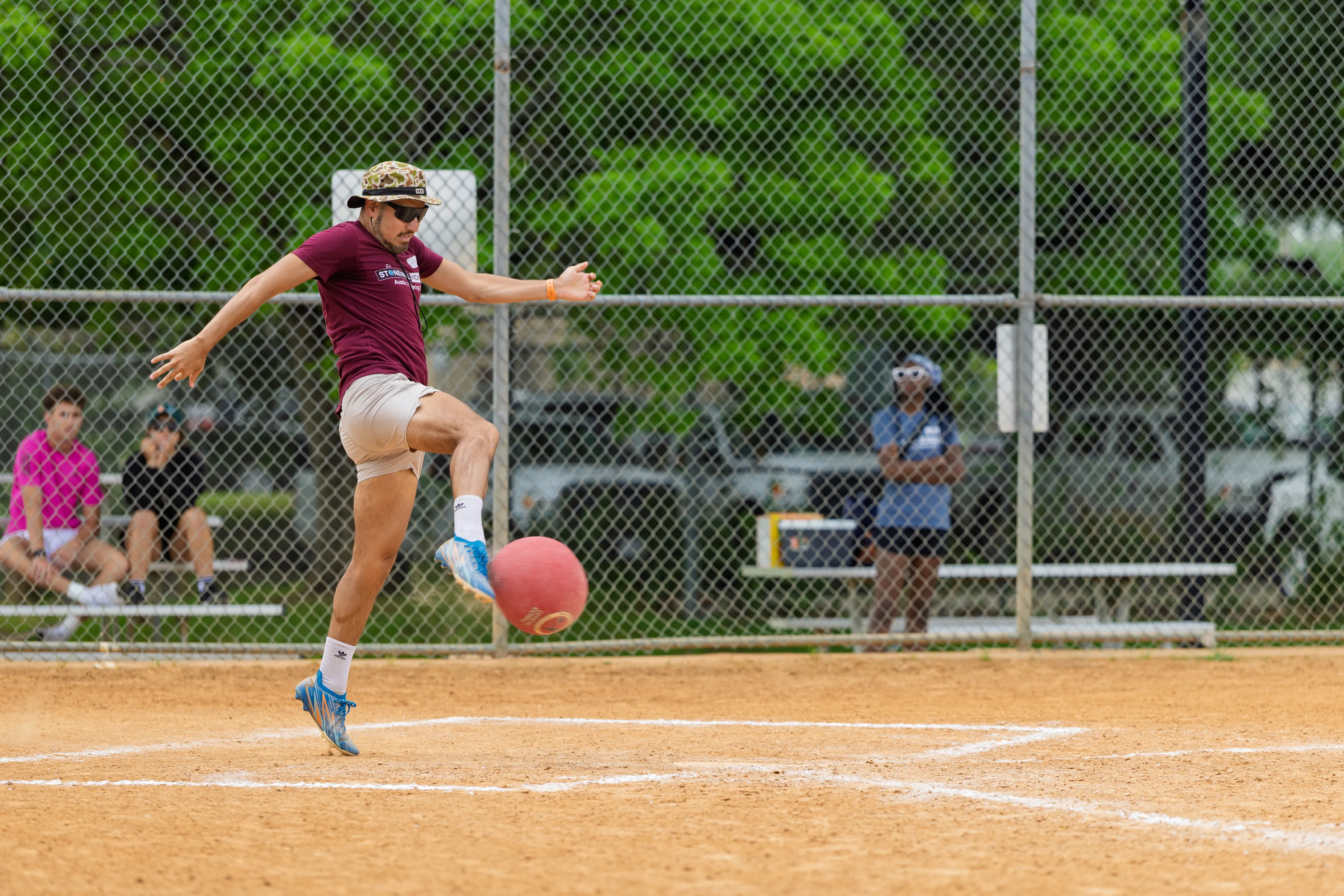 On a sandy kickball field, a player wearing a maroon shirt, light shorts, white socks, blue cleats, a patterned bucket hat, and sunglasses launches a red kickball with a high, airborne kick near home plate. Teammates and spectators watch from benches behind a chain-link fence with green trees beyond.