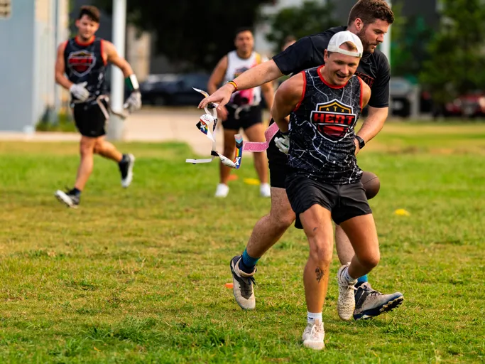 On a grassy flag football field, ATX Heat (Orange) players run drills as a defender yanks a runner’s flag mid-sprint; athletes wear black ATX Heat jerseys with orange trim, flag belts, shorts, cleats, and a backward cap, with cones marking the practice area.