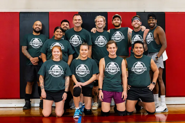 TPSL Dodgeball Summer 2025 team photo: a group of players in matching dark green “Texas Premier” shirts poses on an indoor dodgeball gym floor, some kneeling in front and others standing with arms around teammates. They wear athletic shorts and sneakers, with a few knee pads, headbands, and a cap visible, set against red-and-black wall padding.