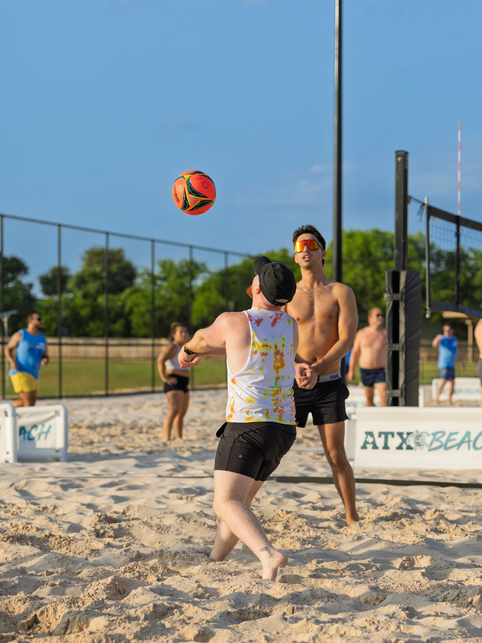 Two sand volleyball players are focused on the ball, with one preparing to bump it while wearing a tie-dye tank top, shorts, and a cap; the game takes place on a sunny sand court surrounded by a fence, with other players and a net visible in the background.