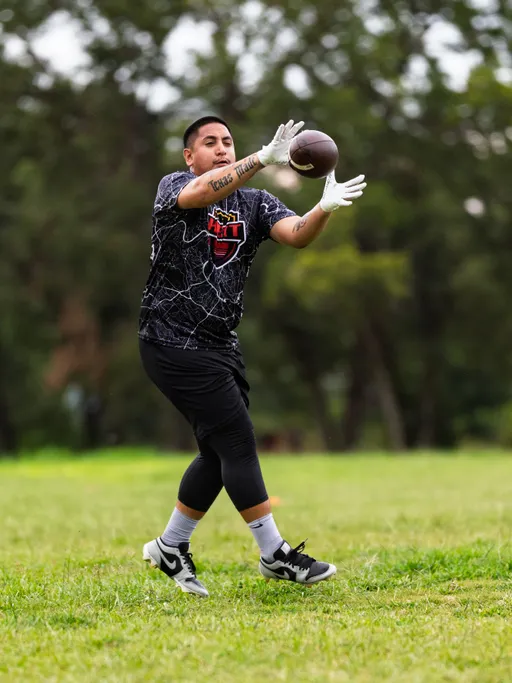 An ATX Heat athlete in a black patterned team jersey with an orange logo, white receiver gloves, leggings, and cleats stretches forward to catch a football during flag football practice for Gay Bowl 2025 on a grassy field, with trees blurred in the background.