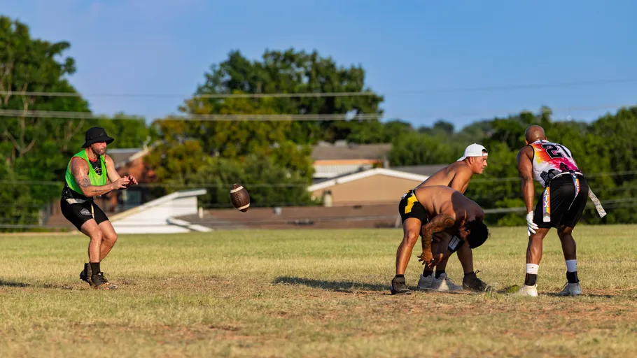 On a sunny grassy field, ATX Heat (Black) players practice flag football: a center snaps the ball to a quarterback in a bright green pinnie and bucket hat as two defenders with flag belts—one in a pink “21” jersey and gloves—prepare to rush, with houses and trees in the background.