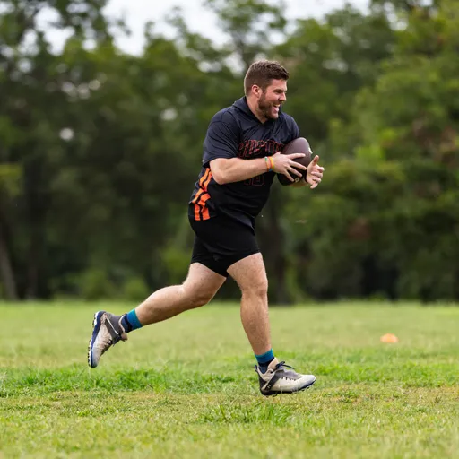 An ATX Heat (Orange) player sprints across a grassy field clutching a football, wearing a black jersey with orange “HEAT” graphics, black shorts, cleats, and bright socks. Orange cones mark the flag football practice area with trees in the background.