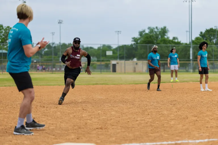 A kickball player in a maroon shirt and black athletic gear is running towards a base while several players in teal shirts watch from the field on a sandy outdoor kickball field.