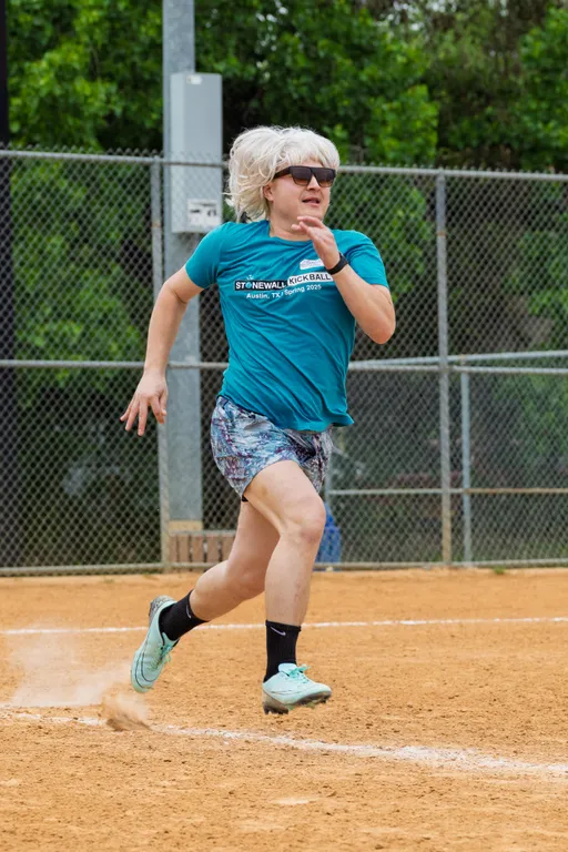 A player is running on a kickball field, wearing a teal "Stonewall Kickball - Austin, TX - Spring 2025" shirt, patterned shorts, light-colored athletic shoes, black socks, sunglasses, and a light wig. The background shows a chain-link fence and greenery.