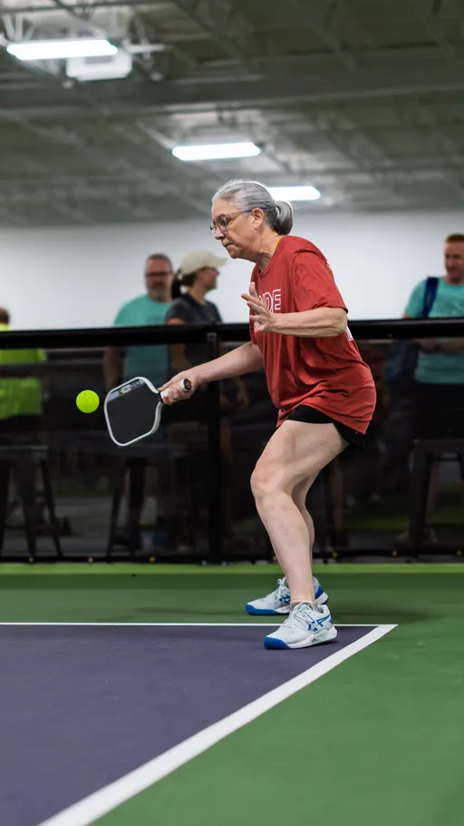 A pickleball player in athletic apparel prepares to hit a ball with a paddle on an indoor court, while spectators watch from behind the black barrier.