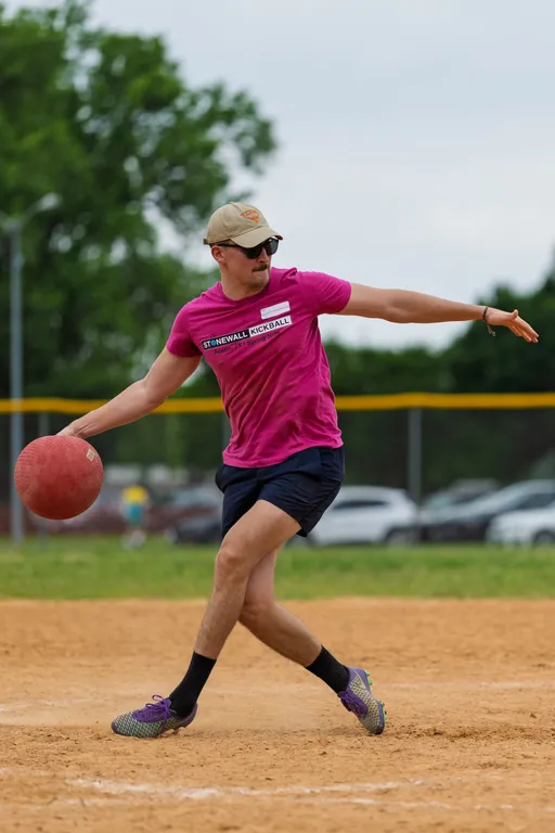 A player wearing a bright pink "Stonewall Kickball" shirt, shorts, a cap, and sunglasses is in the process of pitching a red kickball on a dirt field, with trees and a fence in the background.